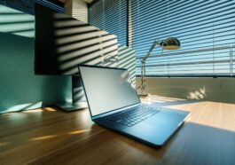 A modern workspace featuring a laptop, desk lamp, and a large monitor by a window with blinds.