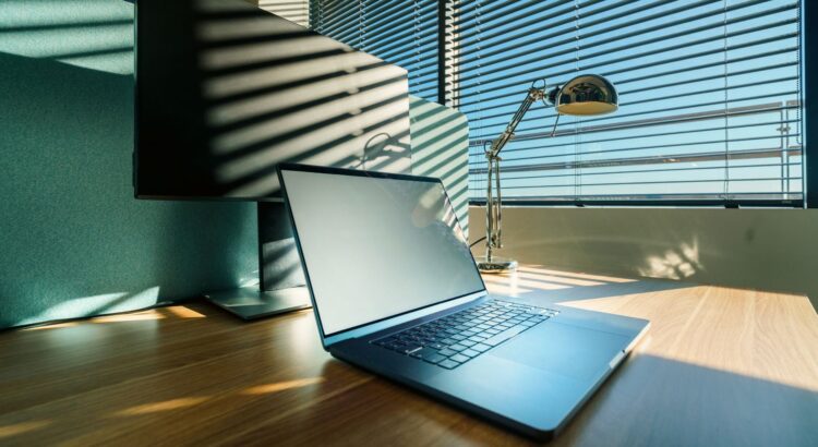 A modern workspace featuring a laptop, desk lamp, and a large monitor by a window with blinds.