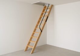 A wooden loft ladder leading up to a ceiling hatch in a minimalist room